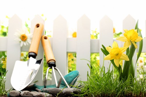 A close-up view of a small garden scene showing two gardening toolsâ€”a trowel and a hand forkâ€”resting on a patch of dark soil next to bright yellow daffodils and white daisies with yellow centers, set against a white picket fence background. The garden features a lush, green grass lawn, with the tools positioned in the foreground, indicating recent or ongoing gardening activity. The scene is illuminated by natural sunlight, creating a cheerful and inviting outdoor environment typical of a well-maintained private garden in Bayswater or surrounding areas. The overall composition emphasizes garden maintenance elements and seasonal flower growth, aligning with professional gardening and hedge trimming services offered by Hedge Trimming Bayswater, providing visual context for outdoor lawn and planting care in the local area.