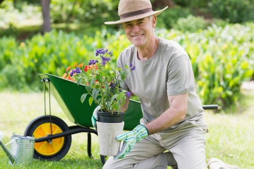 A smiling middle-aged man wearing a wide-brimmed straw hat, grey t-shirt, and gardening gloves is kneeling on a well-maintained lawn in a garden, holding a pot with flowering plants that include purple and red blooms. Behind him, there is a green garden wheelbarrow with a bright yellow wheel and a metal watering can placed nearby on the grass. The garden features a backdrop of lush greenery with trees and shrubs, and a hedge line runs along the background. The scene captures a bright, sunny day with natural light illuminating the outdoor space, emphasizing the vibrant colours of the flowers, the healthy green grass, and the variety of garden elements. This setting reflects professional gardening services such as hedge trimming and general lawn care, suggesting a well-maintained outdoor area typical of residential properties in Bayswater or surrounding areas within the postcode close to London, demonstrating a neat and appealing landscape.